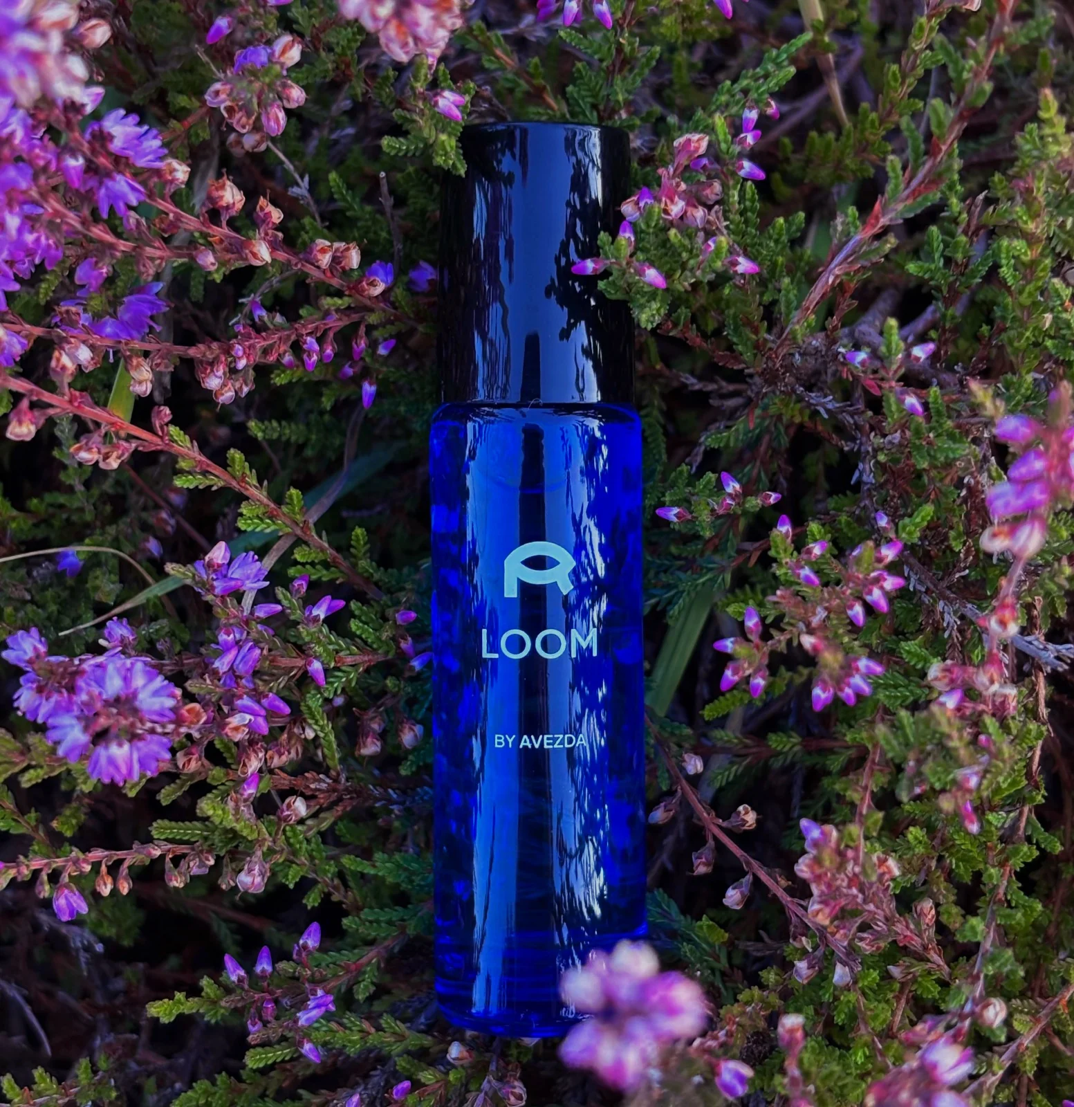 Close-up of the blue Loom Eye Serum bottle surrounded by purple flowers
